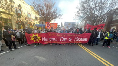 Marchers hold a read and yellow banner that reads, "In the spirit of Metacom - National Day of Mourning." It is as wide as the whole street.